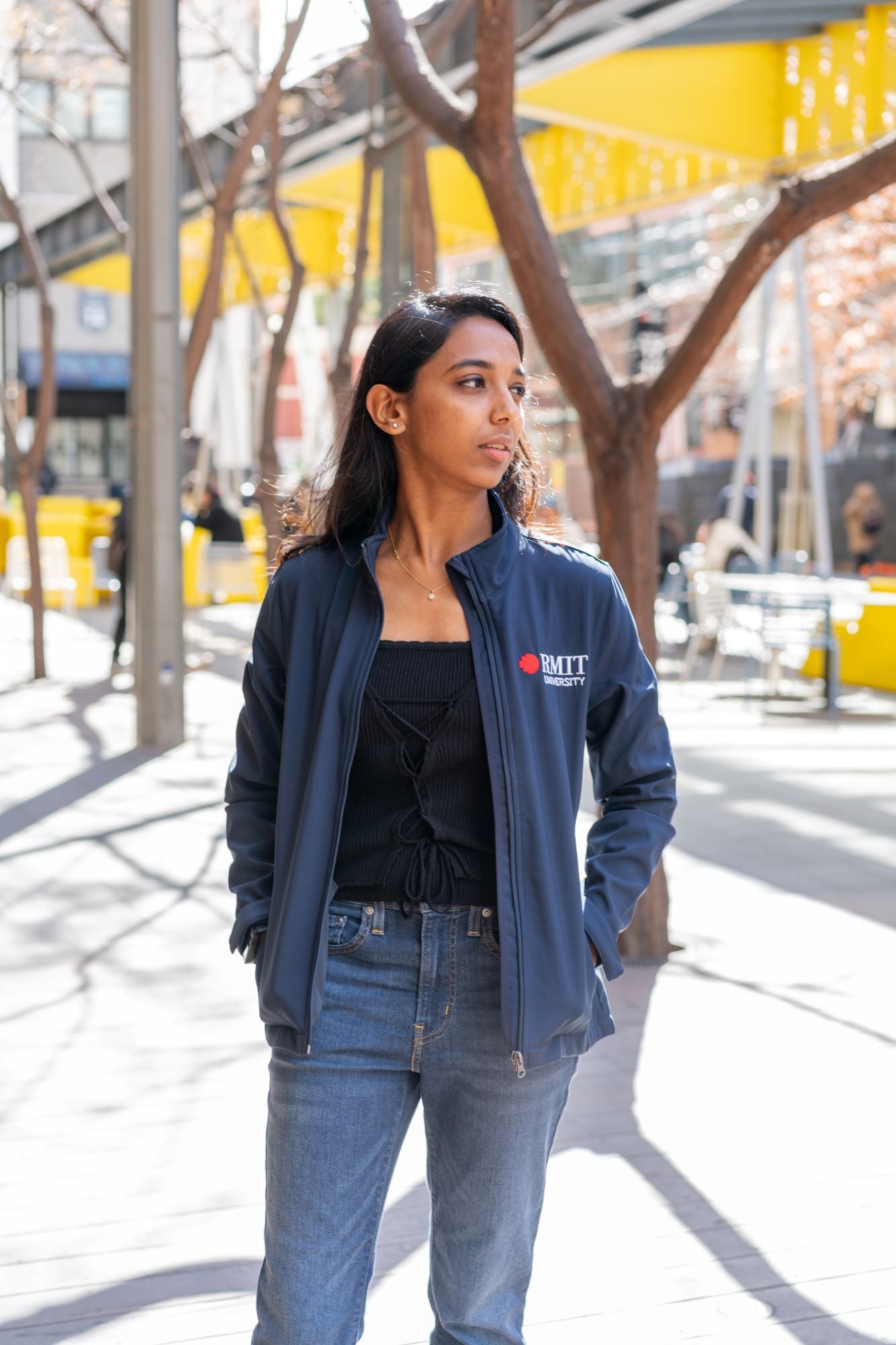 Full front view of the RMIT Softshell Jacket in navy, worn by a female model standing in an urban outdoor setting. The model is looking to her left with the jacket unzipped, revealing a black top underneath. She pairs the jacket with mid-wash blue denim jeans. The RMIT logo is clearly embroidered on the left chest. The jacket features a high collar, structured fit, and long sleeves, styled casually for everyday wear.
